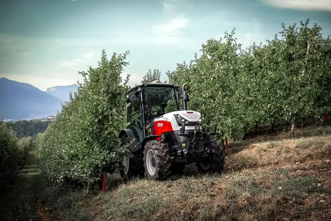 STEYR Spezial Obstbautraktor an einem Hang zwischen Apfelbäumen mit Bergen im Hintergrund
