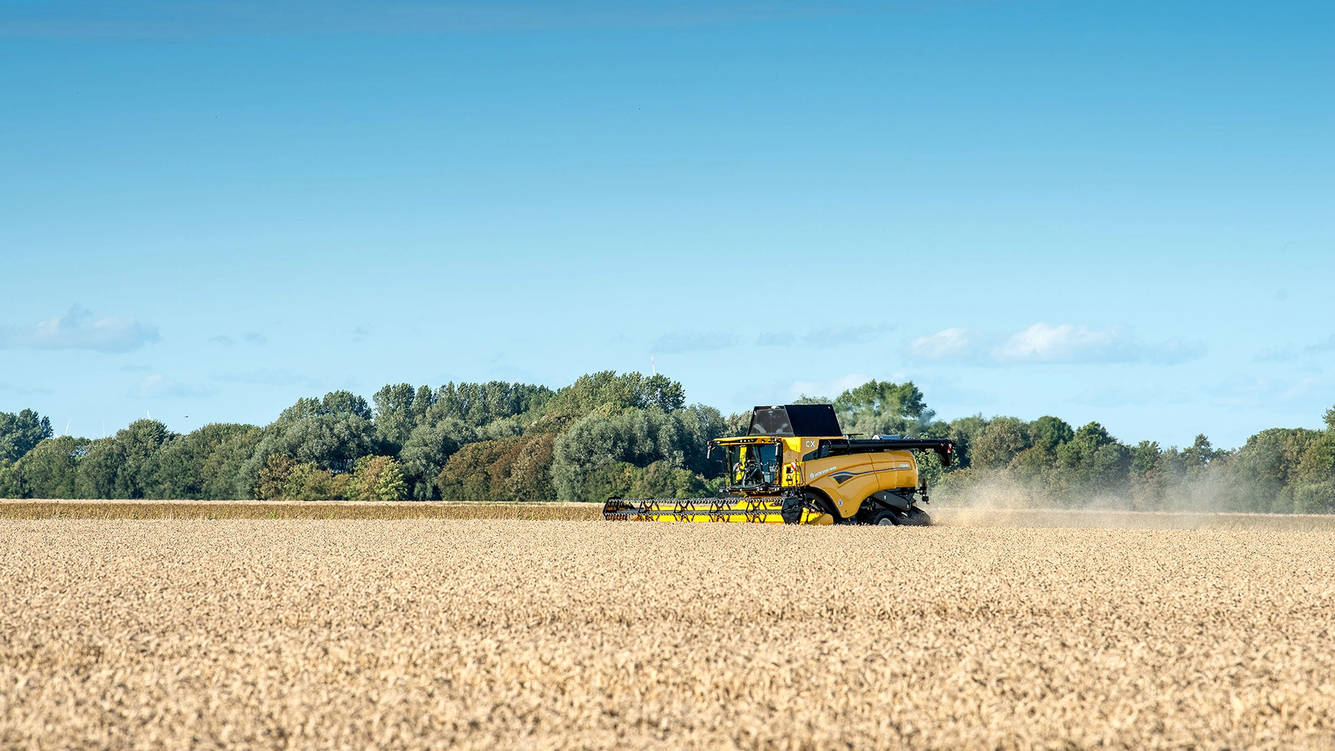 New Holland CX7 & CX8 combine harvester models side by side in a golden field with a picturesque countryside backdrop featuring wind turbines and clear skies.