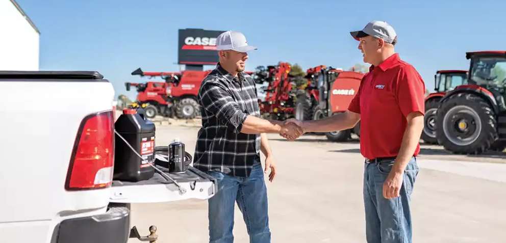 Two men shaking hands beside a white pickup truck with oil containers, with red CASE IH tractors in the background on a sunny day.