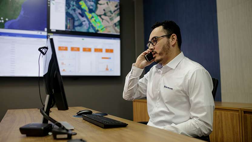 Especialista New Holland em sala de monitoramento atendendo cliente por telefone, com telas exibindo dados agrícolas em tempo real.