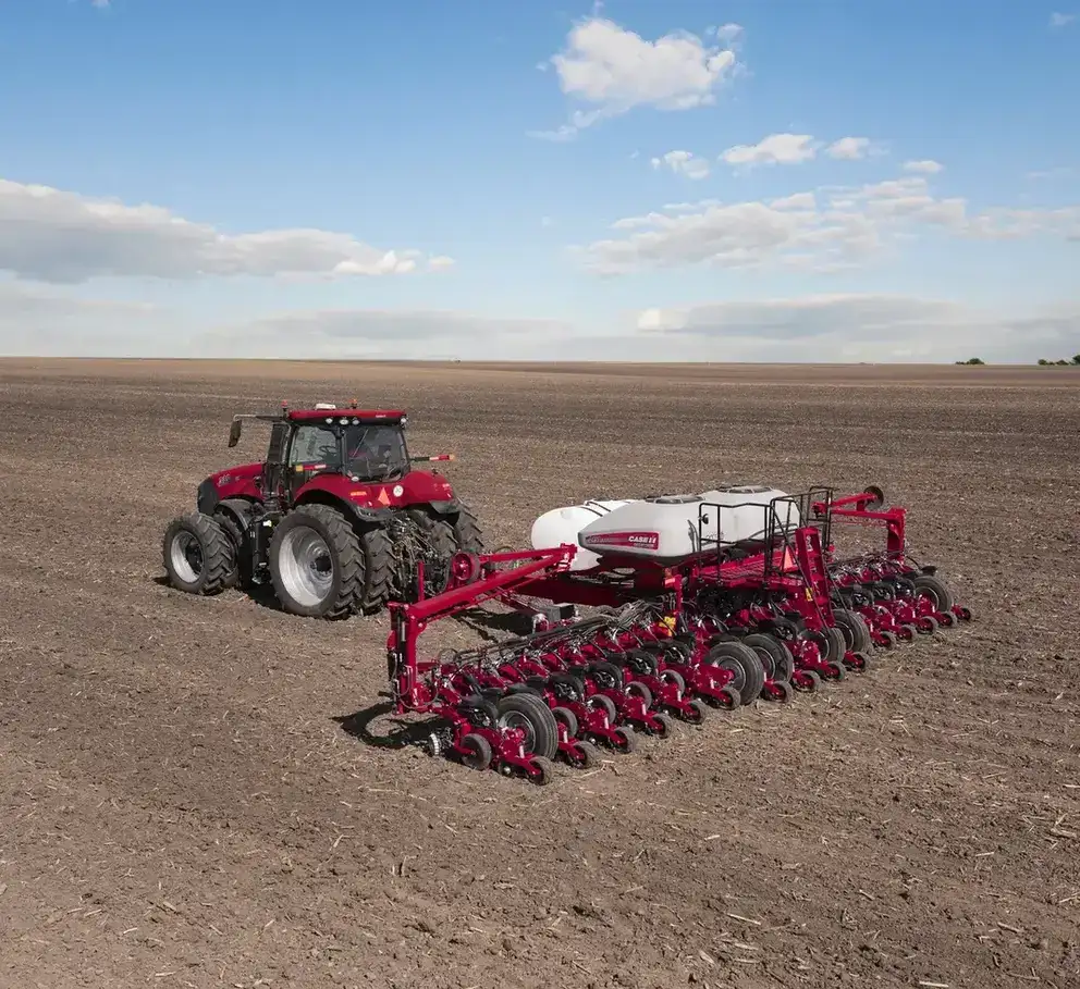 A red Case IH tractor with large tires pulls a white and red planter, traversing a vast, freshly plowed field under a bright blue sky with scattered clouds.