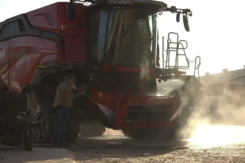 A person power-washes a Case IH combine in the sunlight. Steam rises around the equipment, creating a serene, industrious atmosphere.
