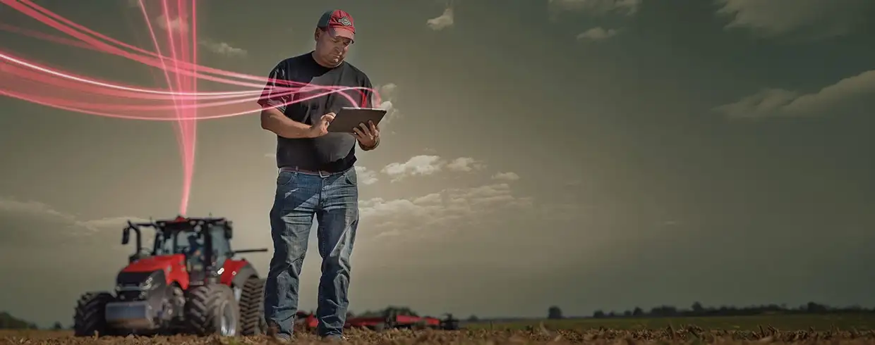 A farmer stands in a field using a tablet, with a Case IH tractor behind him. 