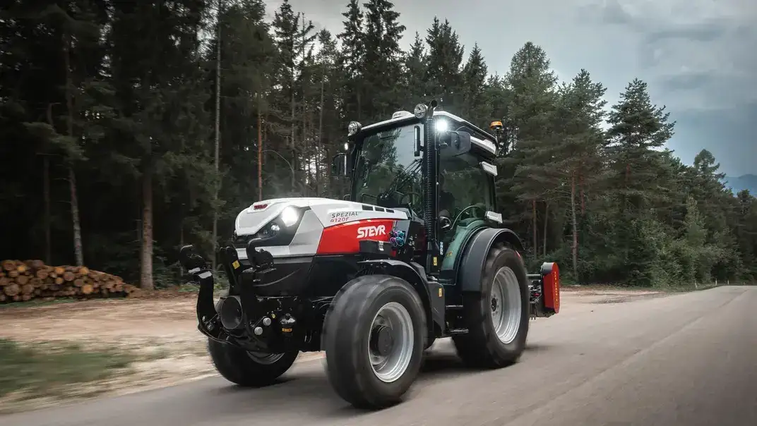 STEYR Spezial 4120F Obstbautraktor auf einer Waldstraße, mit einem Holzstapel am Straßenrand