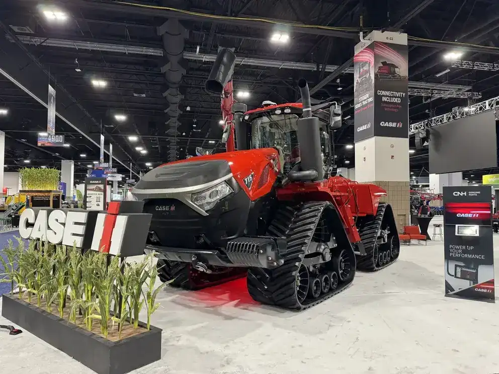 Case IH tractor with large tracks displayed indoors at a trade show. "CASE" sign and corn plants in front. Bright lighting and industrial setting.