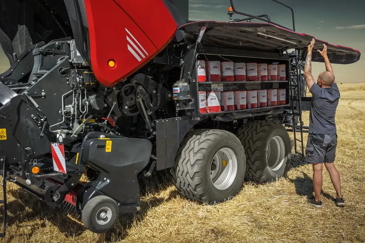 Operator opening the side access panel of a Case IH LB424 XLD large square baler revealing the packer feeder lubricant storage compartment in a field