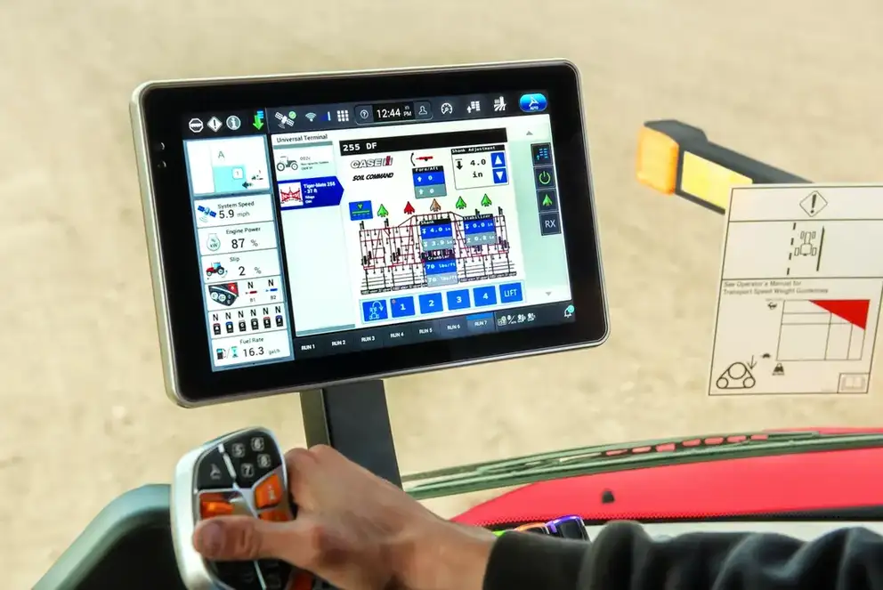 Tablet display shows a precision machine-control map, while a worker holds a joystick; warning diagram and equipment appear nearby.