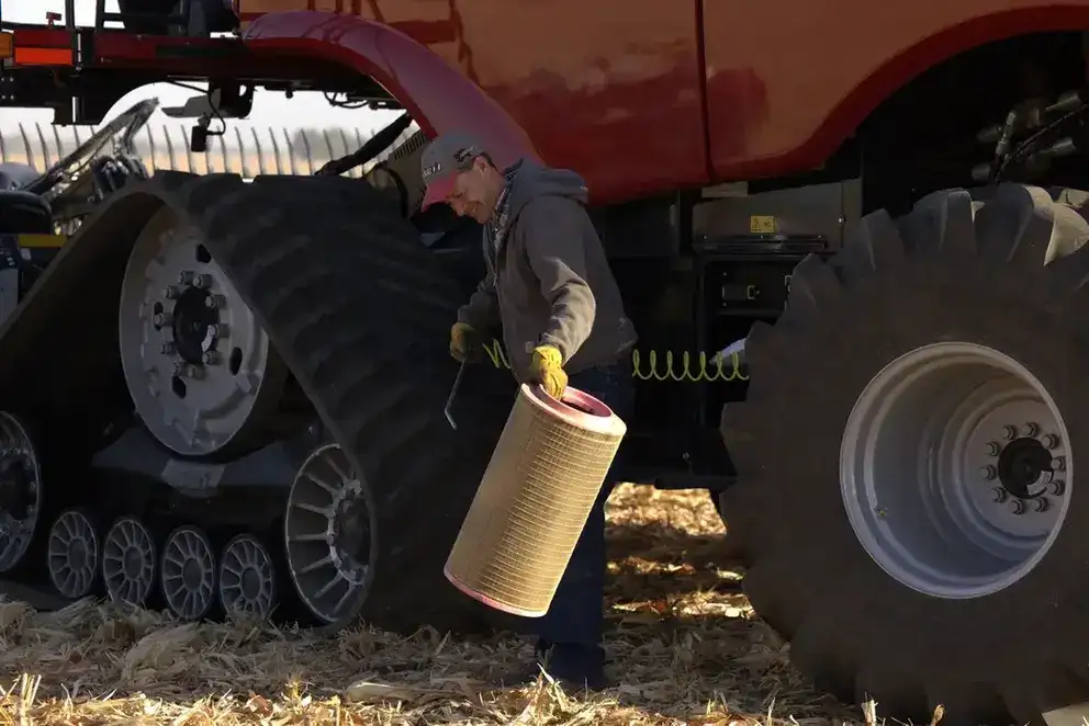 A person wearing a cap and gloves is seen near a large tractor with distinctive treaded wheels, holding a cylindrical object, amidst a field of dry corn stalks.