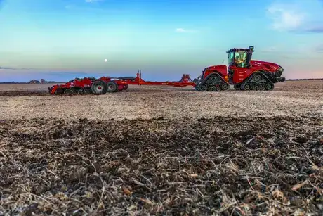 A red Case IH agricultural tractor with tracks working in a plowed field under a blue sky with a visible moon.