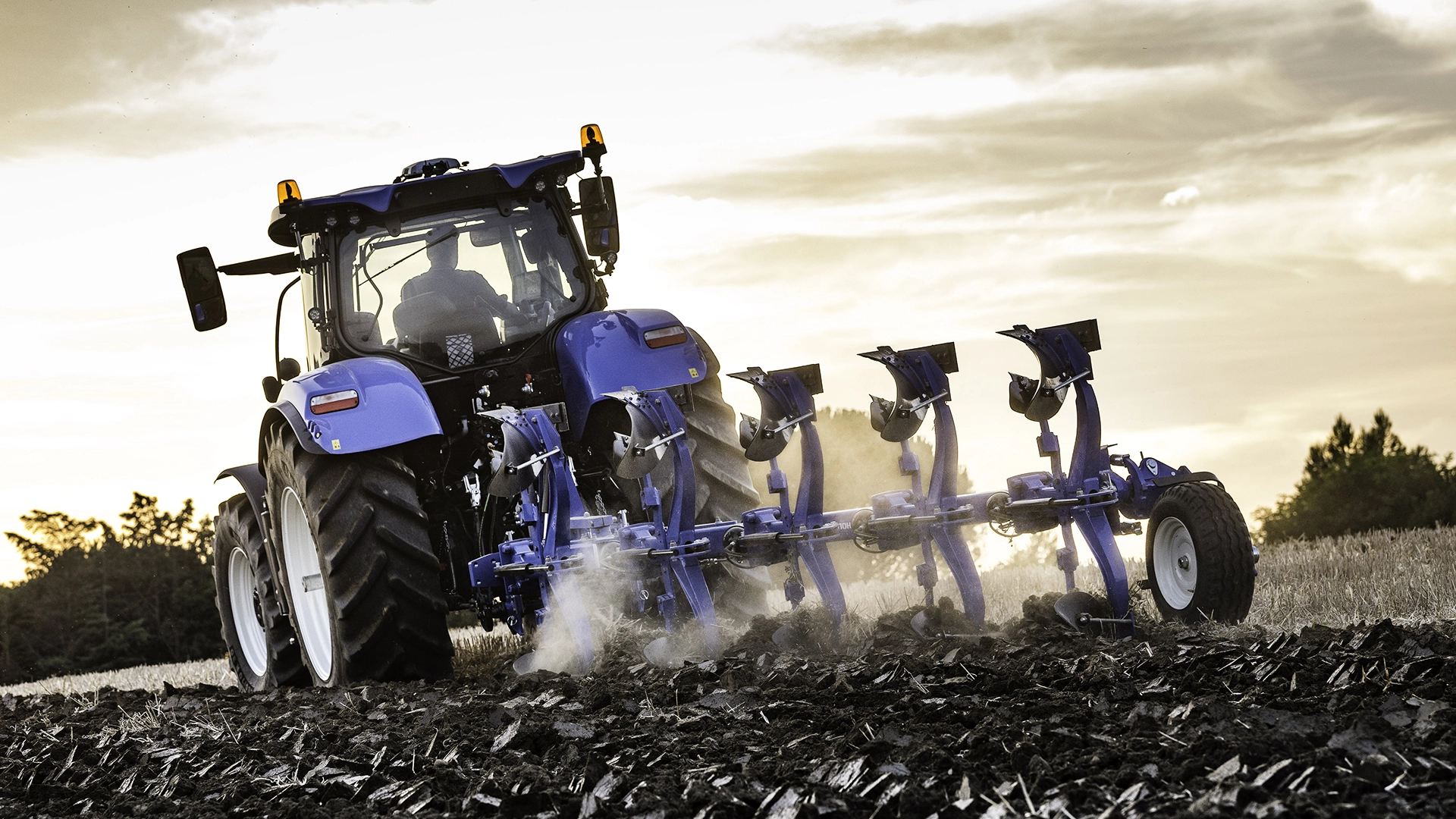 New Holland tractor with a 6-furrow agricultural plough tilling the soil at dusk