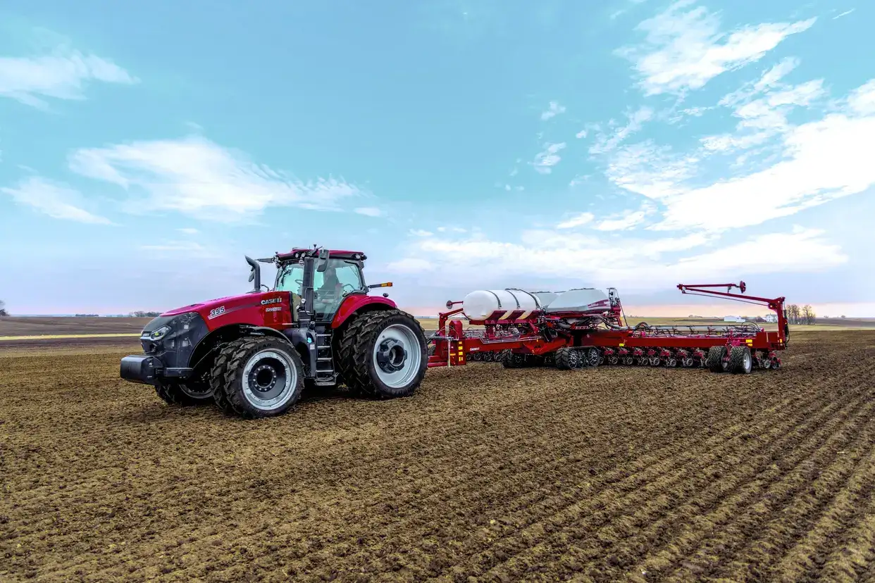 A Case IH tractor with large rear tires pulls a farming implement across a vast, freshly plowed field under a clear blue sky.