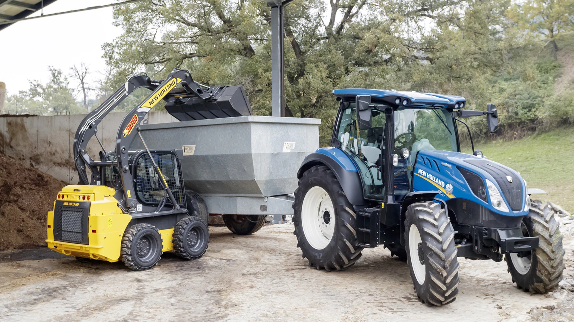 A New Holland skid steer loader is seen transferring material to a gray trailer, while nearby a New Holland blue tractor is parked.