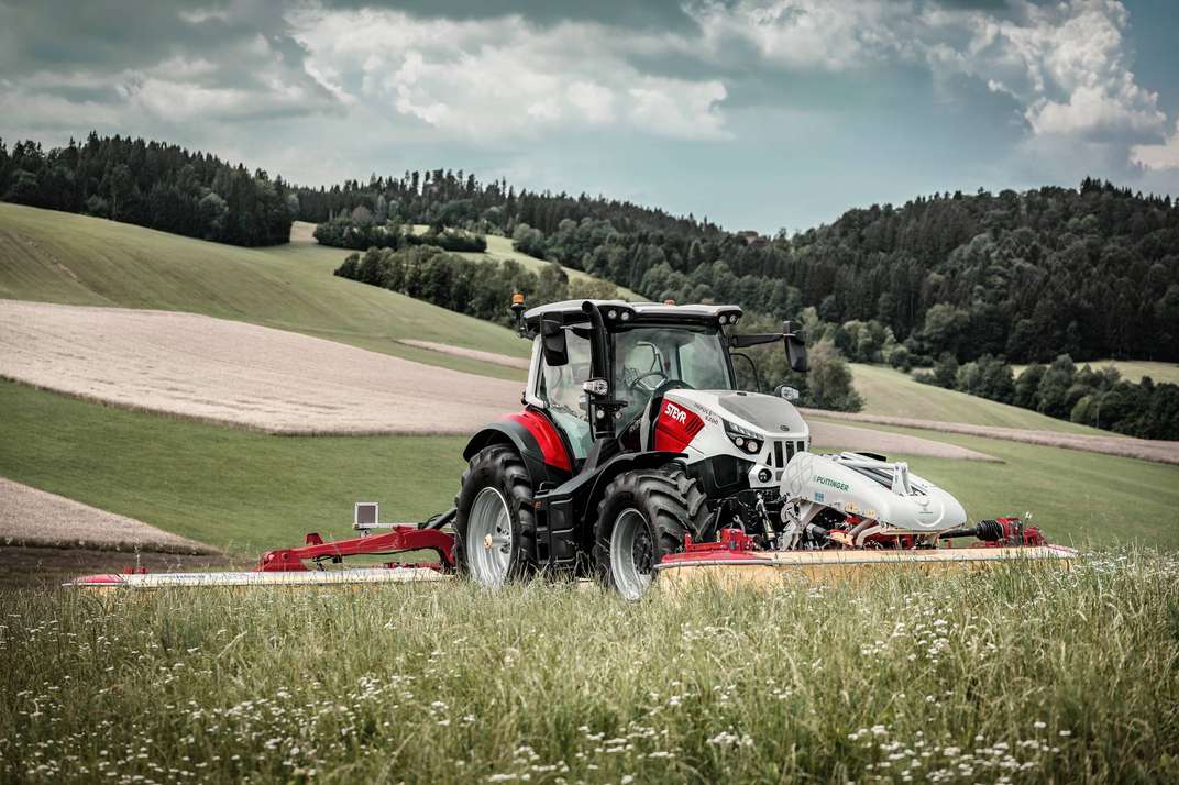 A red and white Steyr Impuls tractor equipped with a front-mounted Pöttinger mower, parked in a lush green meadow with rolling hills and forests in the background under a cloudy sky.