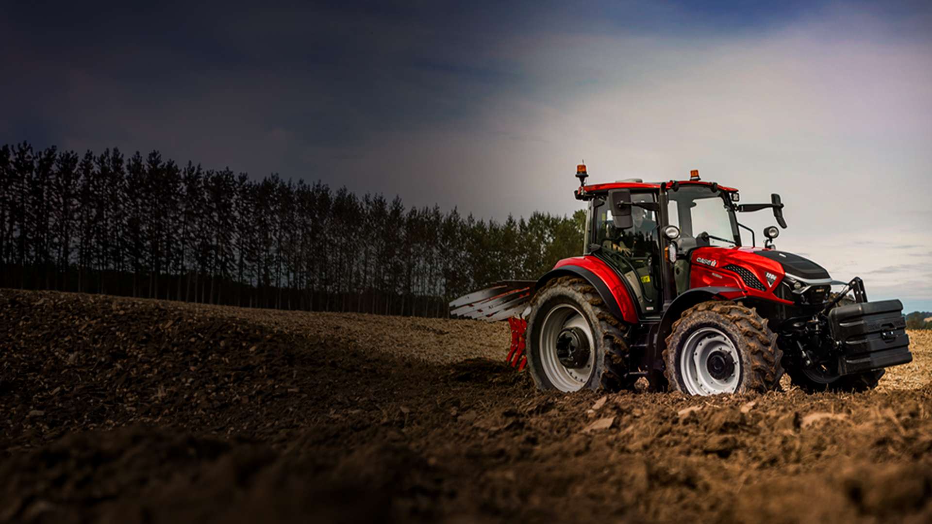 Tractor Farmall 120C rojo trabajando en un campo arado bajo un cielo nublado.