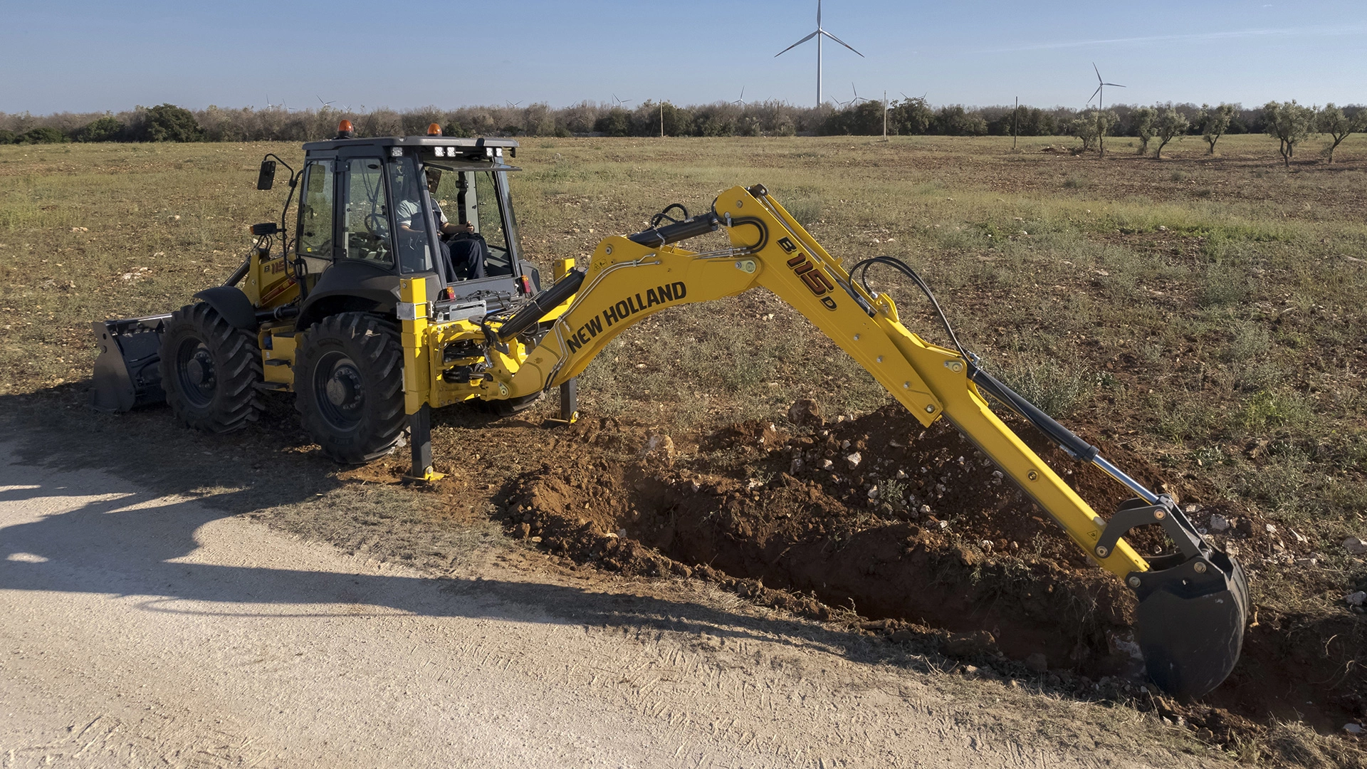 New Holland Backhoe in action, digging up soil, positioned amidst a field with wind turbines in the background.