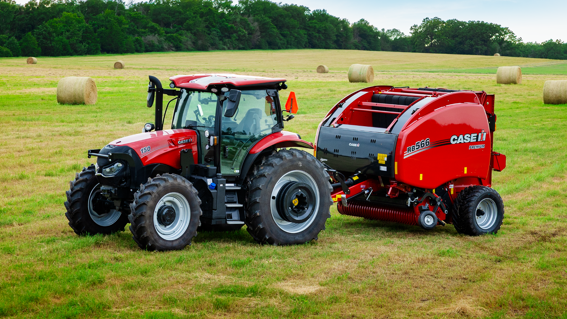 A Case IH tractor with a round hay baler attachment sits in a vast, green field dotted with round hay bales.