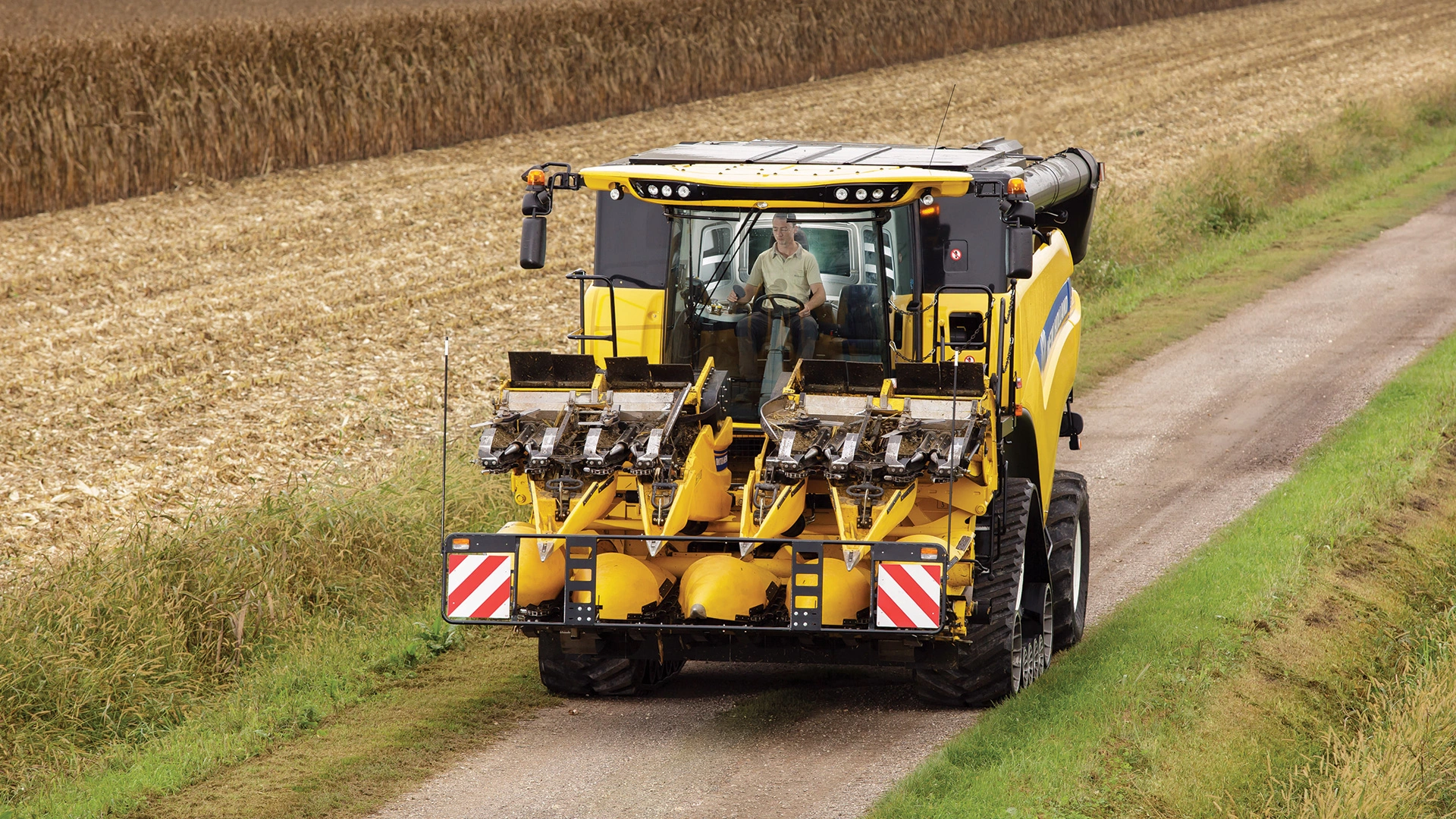 New Holland Maize Headers; yellow harvester on rural road, ready for cornfield harvest with clear skies.