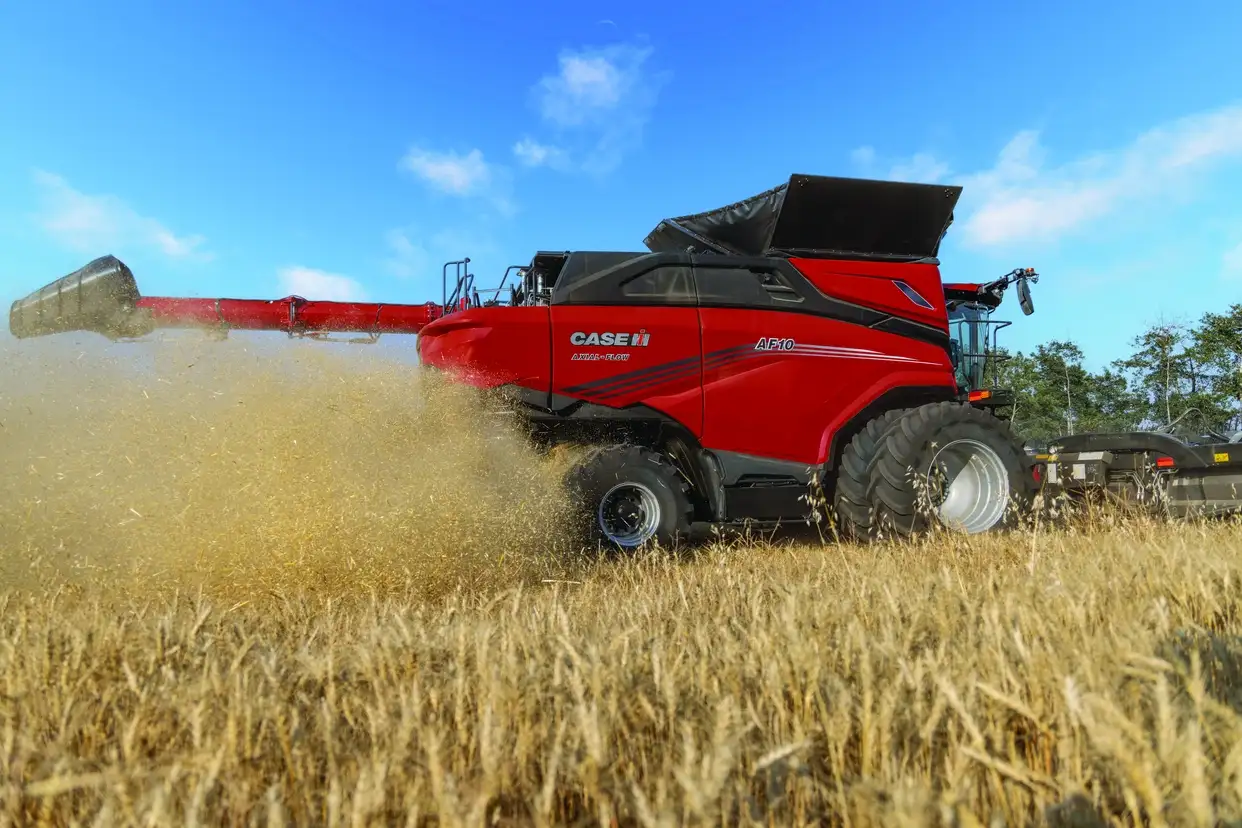 A red Case IH AF10 combine harvester moves through a golden wheat field, kicking up chaff under a bright blue sky with scattered white clouds.