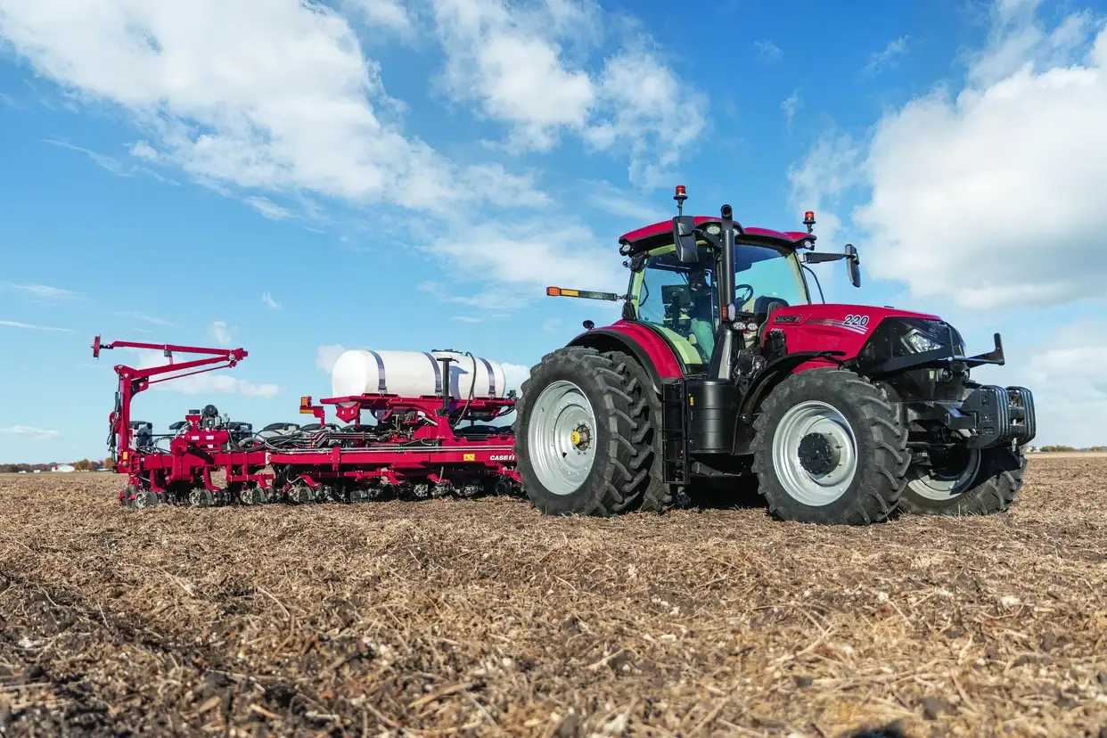 A red Case IH 220 tractor pulling a large agricultural planter with a white tank across a harvested brown field under a bright blue cloudy sky.