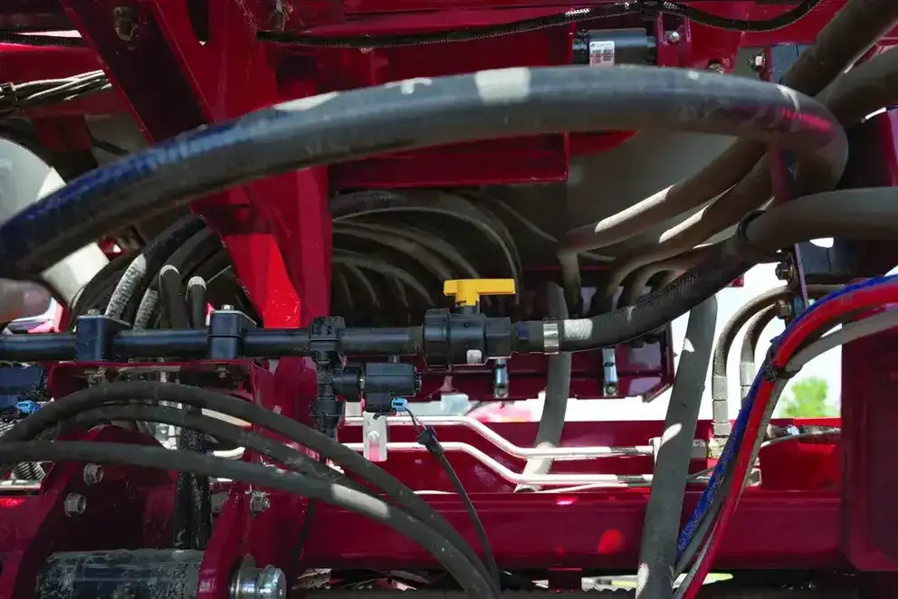 Close-up of industrial machinery with red Case IH metal framework and numerous black hoses and wires. A yellow valve is visible, suggesting complex utility connections. Bright daylight filters through, conveying an industrious atmosphere.