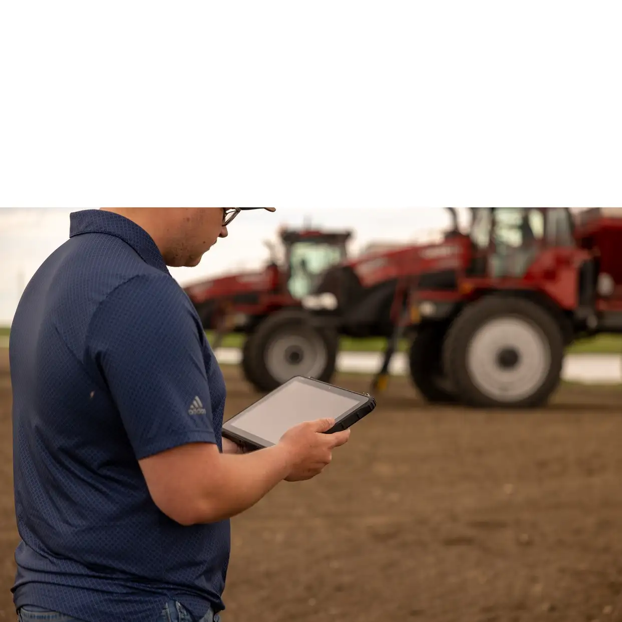 A person in a blue shirt uses a tablet outdoors, with large red farm machinery blurred in the background, conveying a modern agricultural setting.