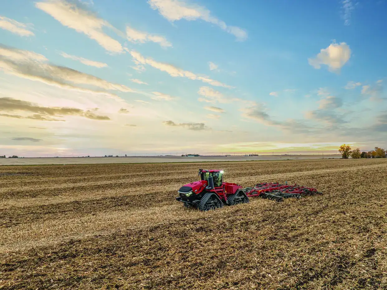 A red Case IH tractor plowing a large field under a colorful sky at sunset, with rows of freshly turned soil.