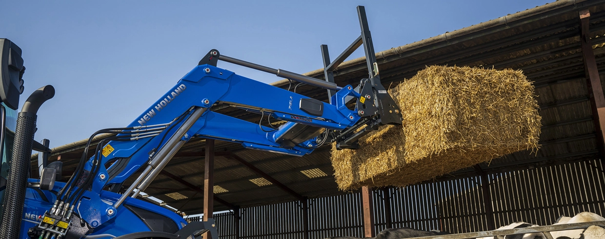 New Holland tractor with a bale fork loader lifting straw in a farm shed.