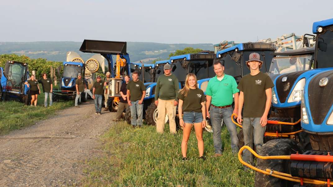 9 people standing in a field in front of New Holland equipment.