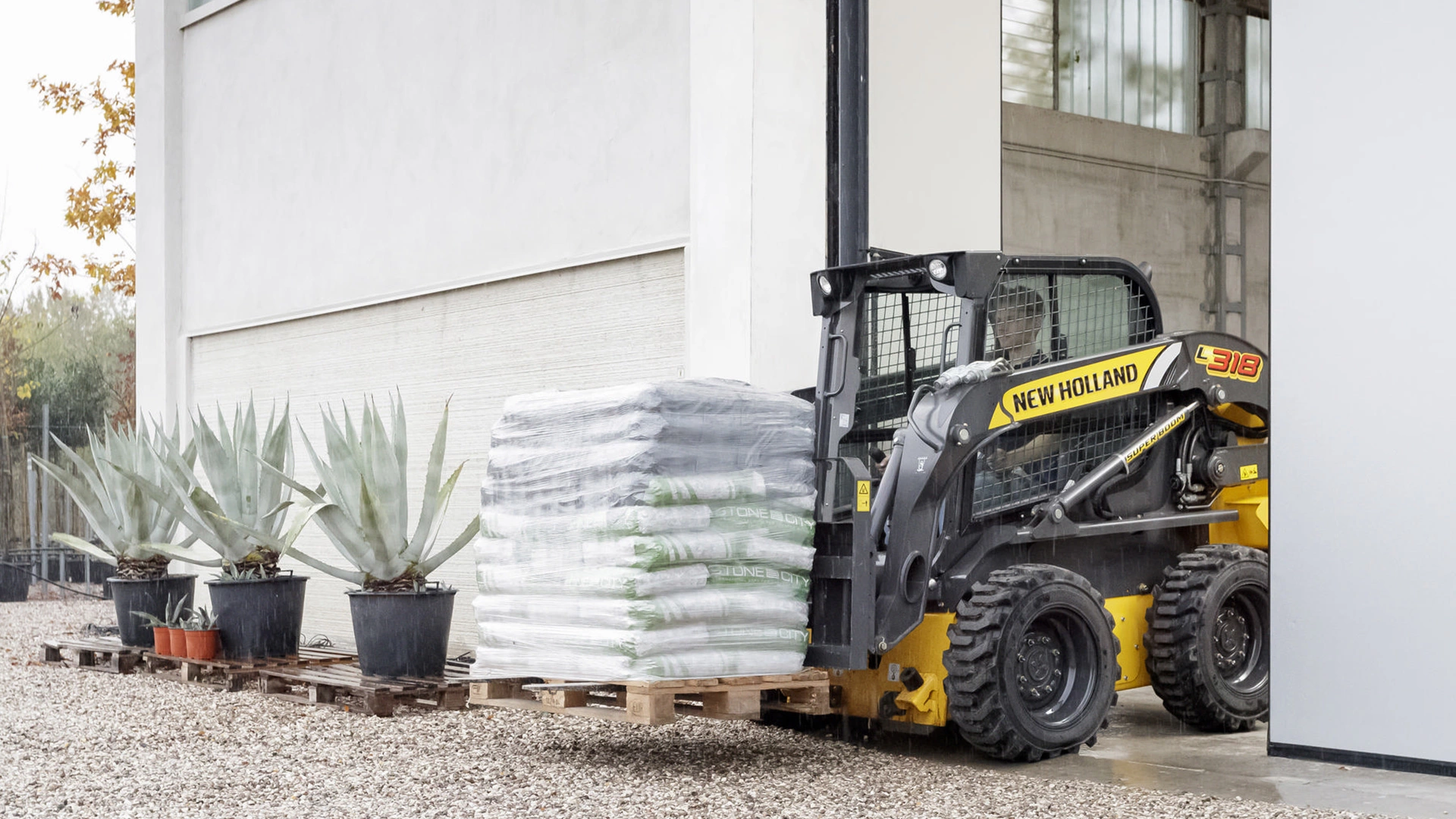 A New Holland skid steer loader in a garden setting, lifting a pallet of soil bags.