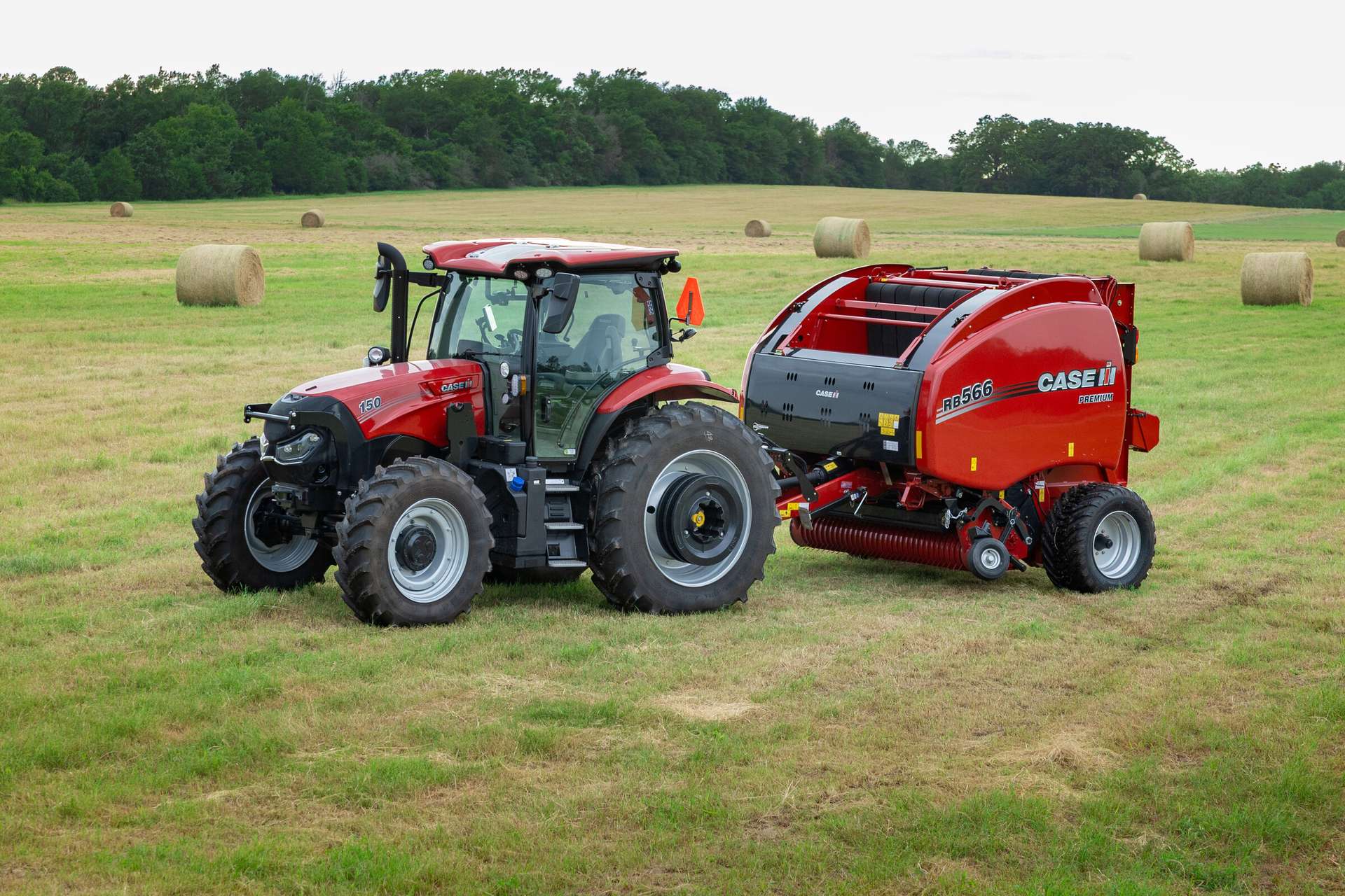 A Case IH tractor with a round hay baler attachment sits in a vast, green field dotted with round hay bales.