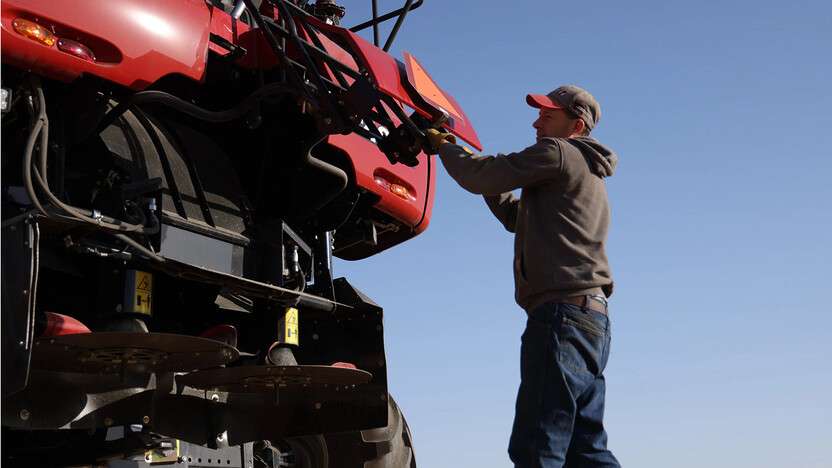 Man in hoodie and cap operating large red agricultural machinery under clear blue sky.