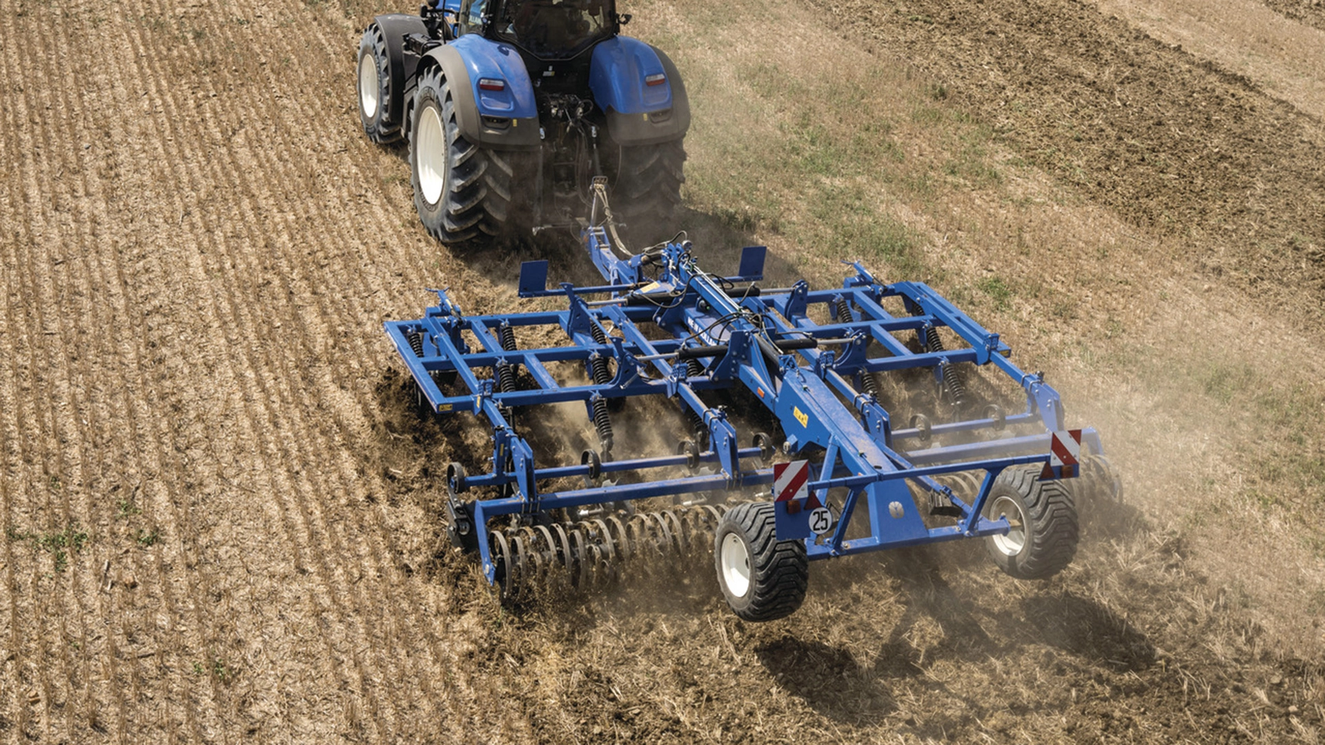 Tractor with mounted stubble cultivator, equipped with rigid tine cultivator and rear rollers, actively working on the field