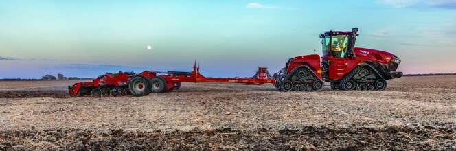A red Case IH agricultural tractor with tracks working in a plowed field under a blue sky with a visible moon.
