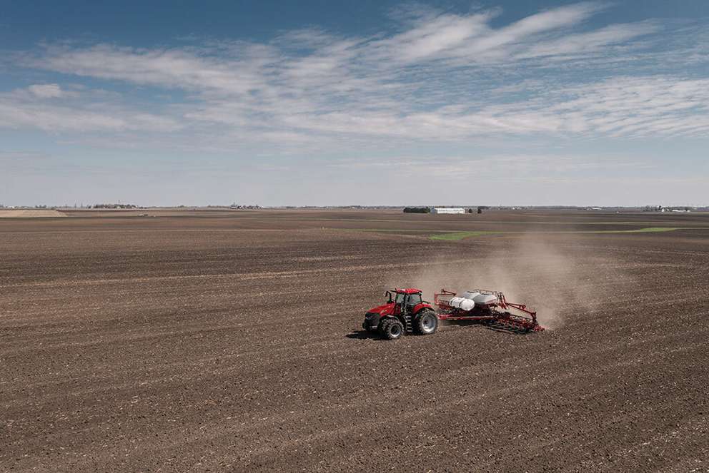 Red Case IH tractor planting in a large open field under a blue sky.