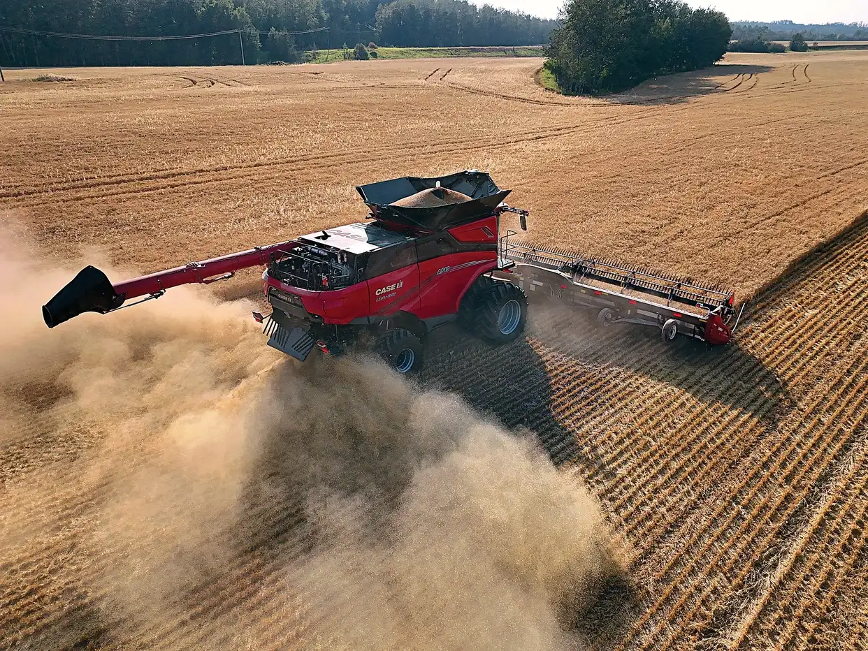 An aerial view of a red Case IH combine harvester moving through a golden wheat field, kicking up a large cloud of dust.