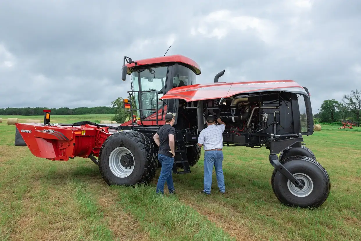 Two farmers inspect a large red Case IH tractor with a mower attachment in a grassy field under cloudy skies, conveying rural farm work.
