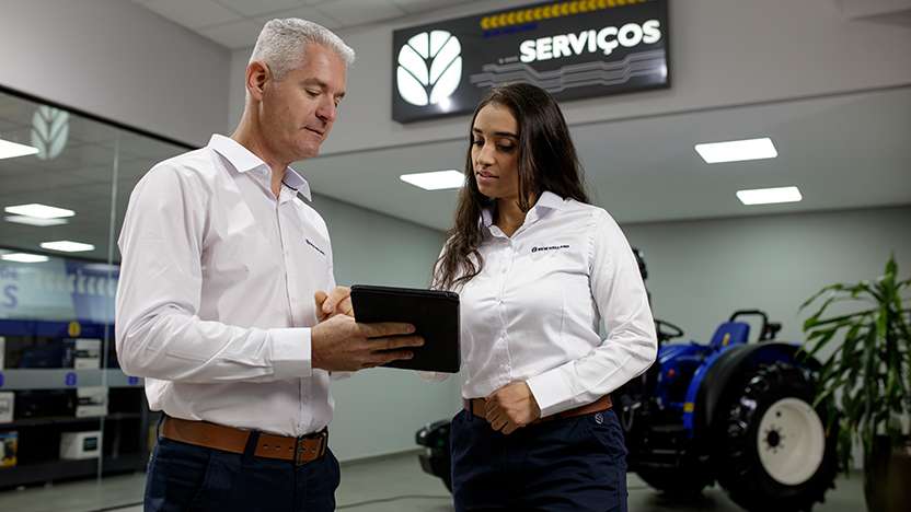 Consultor da New Holland atendendo cliente em concessionária, utilizando tablet para apresentar soluções de serviços personalizados, com trator exposto ao fundo.
