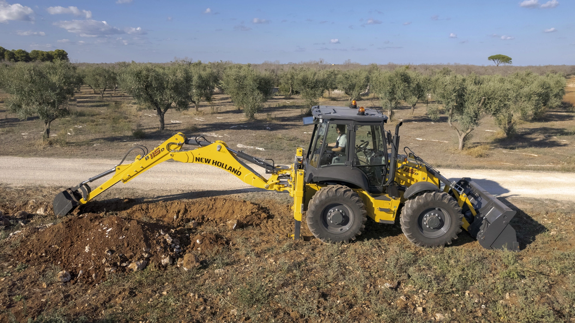 New Holland Backhoe at work, digging in rocky terrain, clear sky, operator in cabin, olive trees in background.