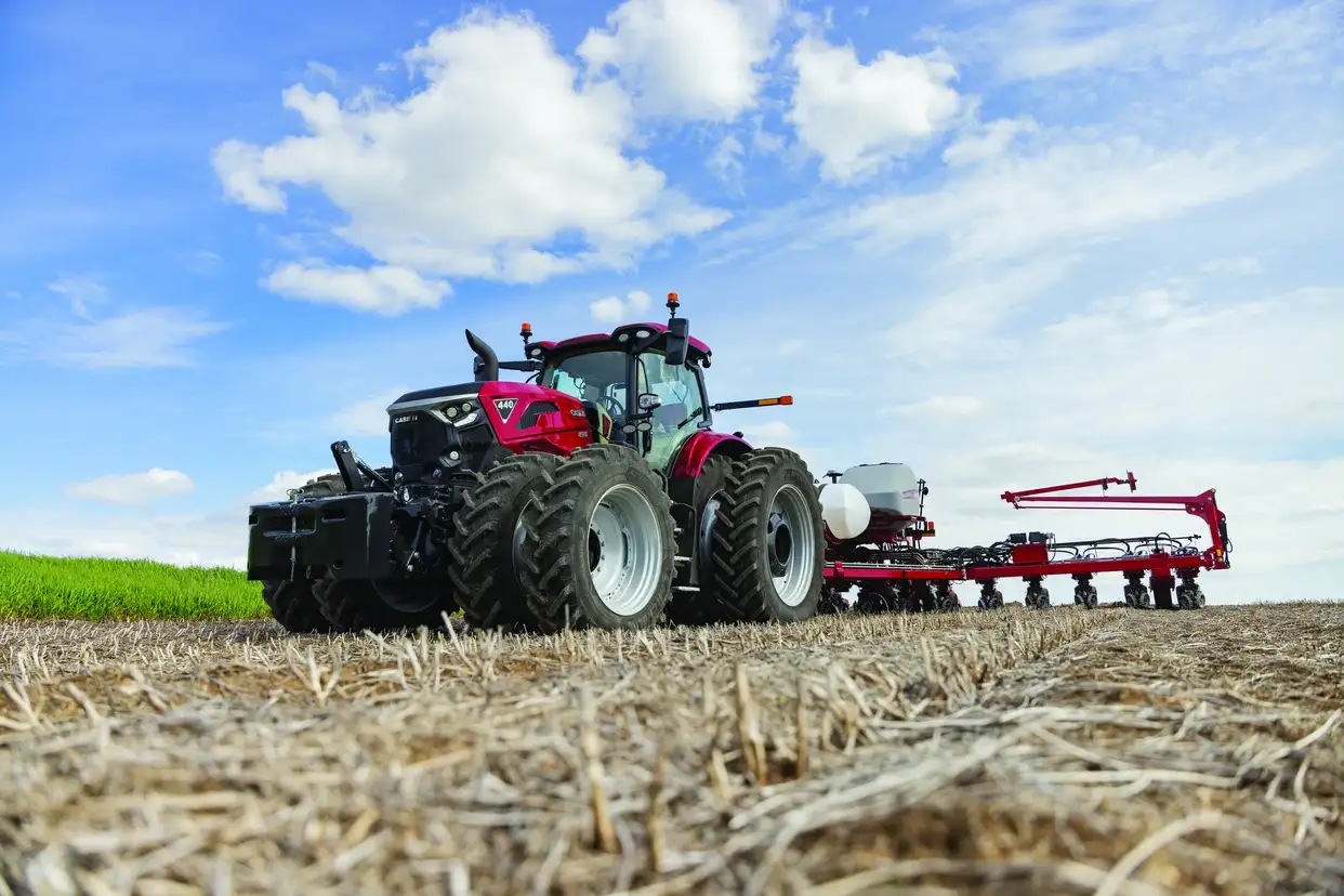 A powerful red Case IH tractor pulls agricultural equipment across a harvested field beneath a bright blue, cloud-filled sky.