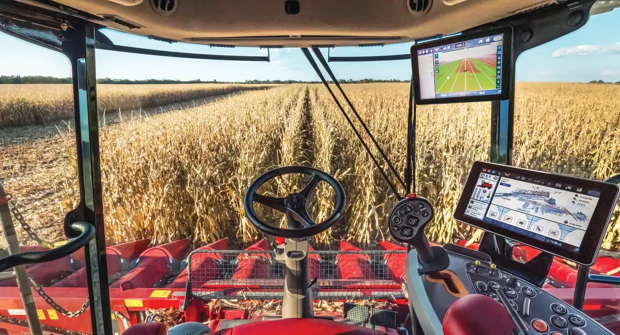 View from a Case IH combine harvester cab over a golden cornfield, with steering wheel, red header, GPS screens, and bright sunny skies.