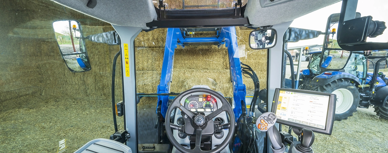View from inside a New Holland tractor cabin with front loader controls and digital display, overlooking a farm.
