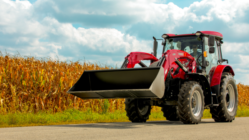 A red CASE IH tractor with an L625 front loader drives along a rural road beside a tall cornfield under a partly cloudy sky, conveying a sense of agricultural work.