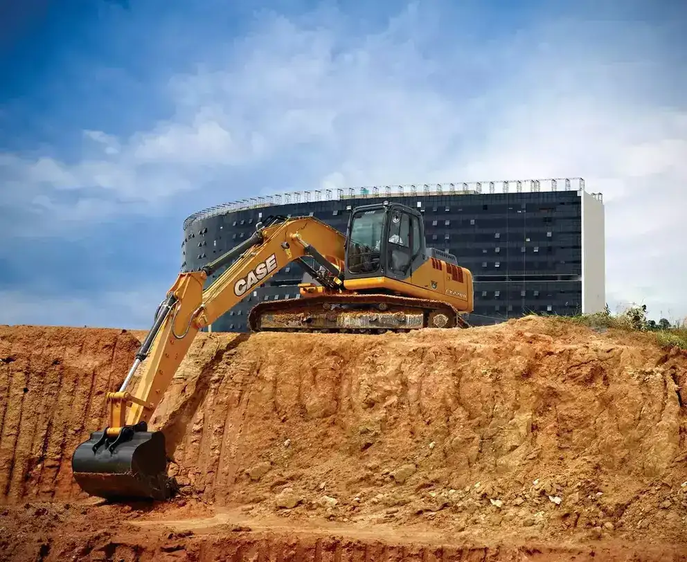 foto ao ar livre da escavadeira hidráulica da Case Construction criando uma superfície estável na terra para preparação do terreno antes da construção. No fundo vemos um prédio elaborado por Oscar Niemeyer.