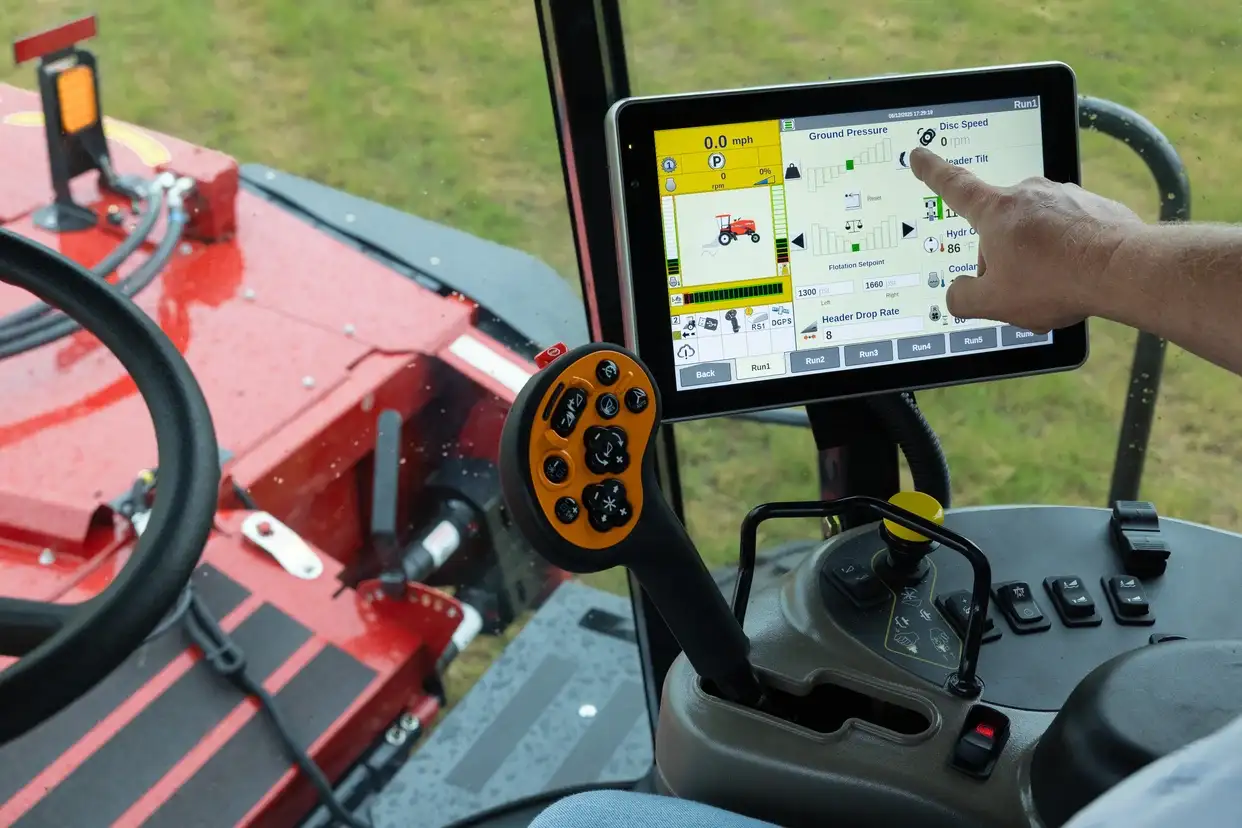 A Case IH tractor operator taps a touchscreen control panel beside an orange joystick; red agricultural mowing equipment is visible outside the cab.