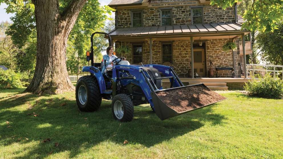 Woman operates a blue tractor in front of a house.