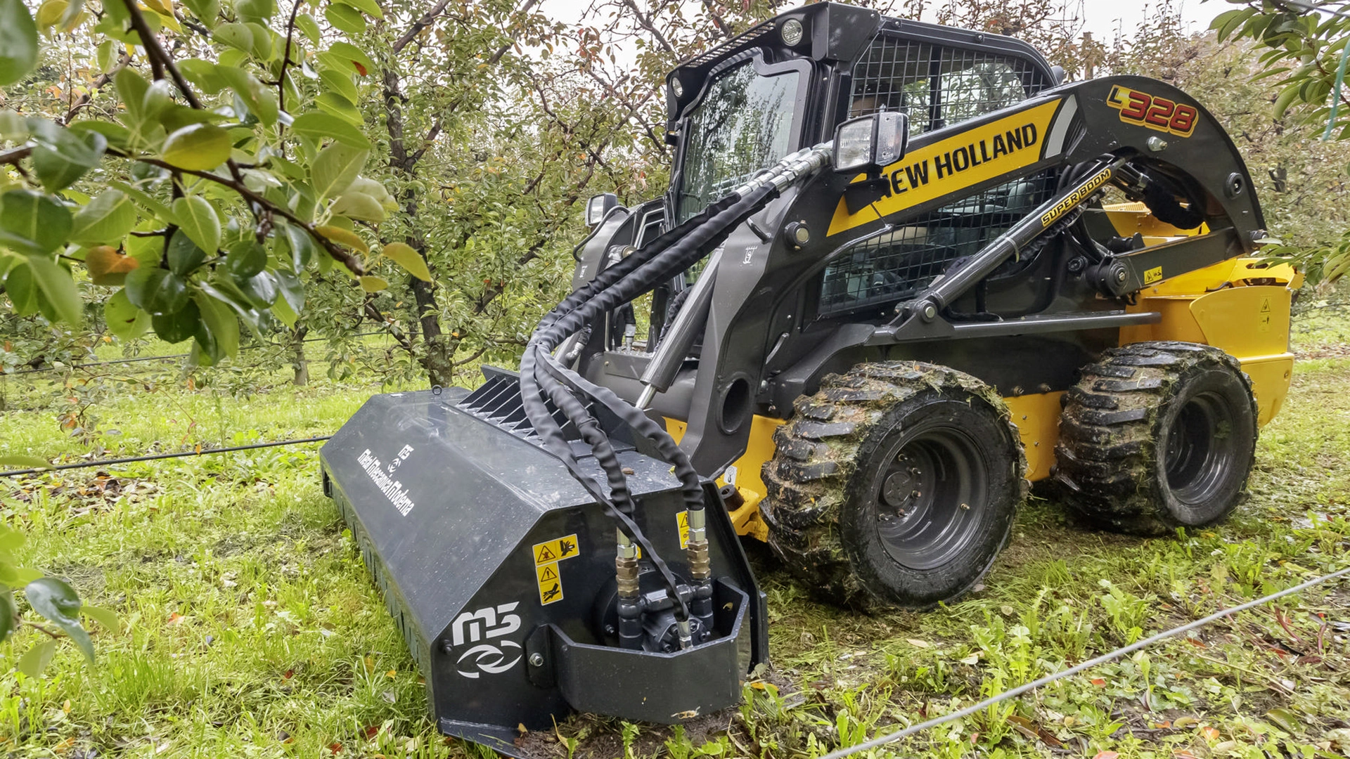 New Holland skid steer loader working in an orchard, muddy tires, amidst apple trees and green grass.