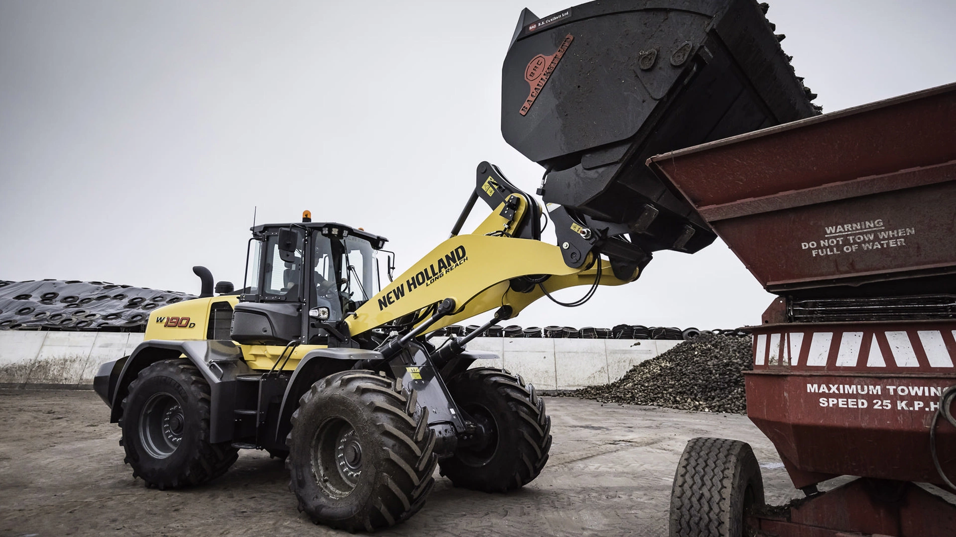 A robust New Holland W190D wheel loader in action, unloading gravel into a red dump trailer, set against an industrial backdrop.