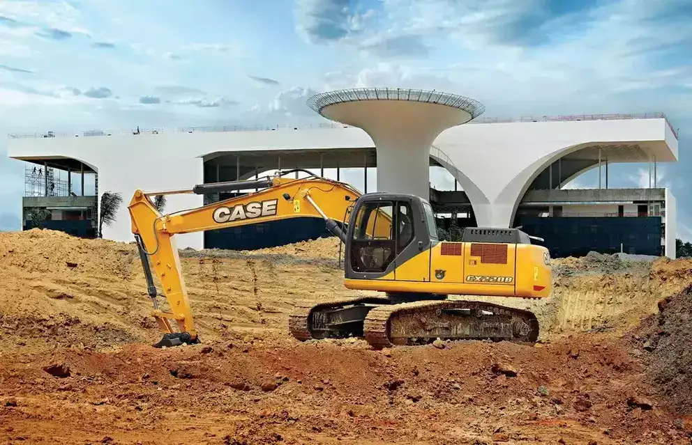 foto ao ar livre da escavadeira hidráulica da Case Construction realizando uma escavação que envolve a remoção de terra. Ao fundo, vemos uma grande construção elaborada por Oscar Niemeyer sendo montada.
