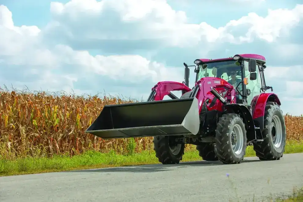 A red CASE IH tractor with an L625 front loader drives along a rural road beside a tall cornfield under a partly cloudy sky, conveying a sense of agricultural work.