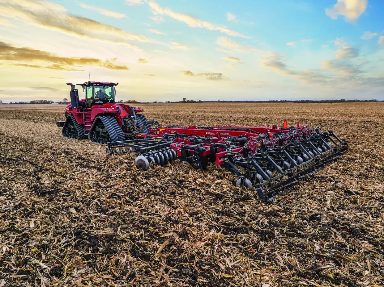 A red Case IH tractor with a tiller attachment working on a harvested cornfield under a cloudy sky.